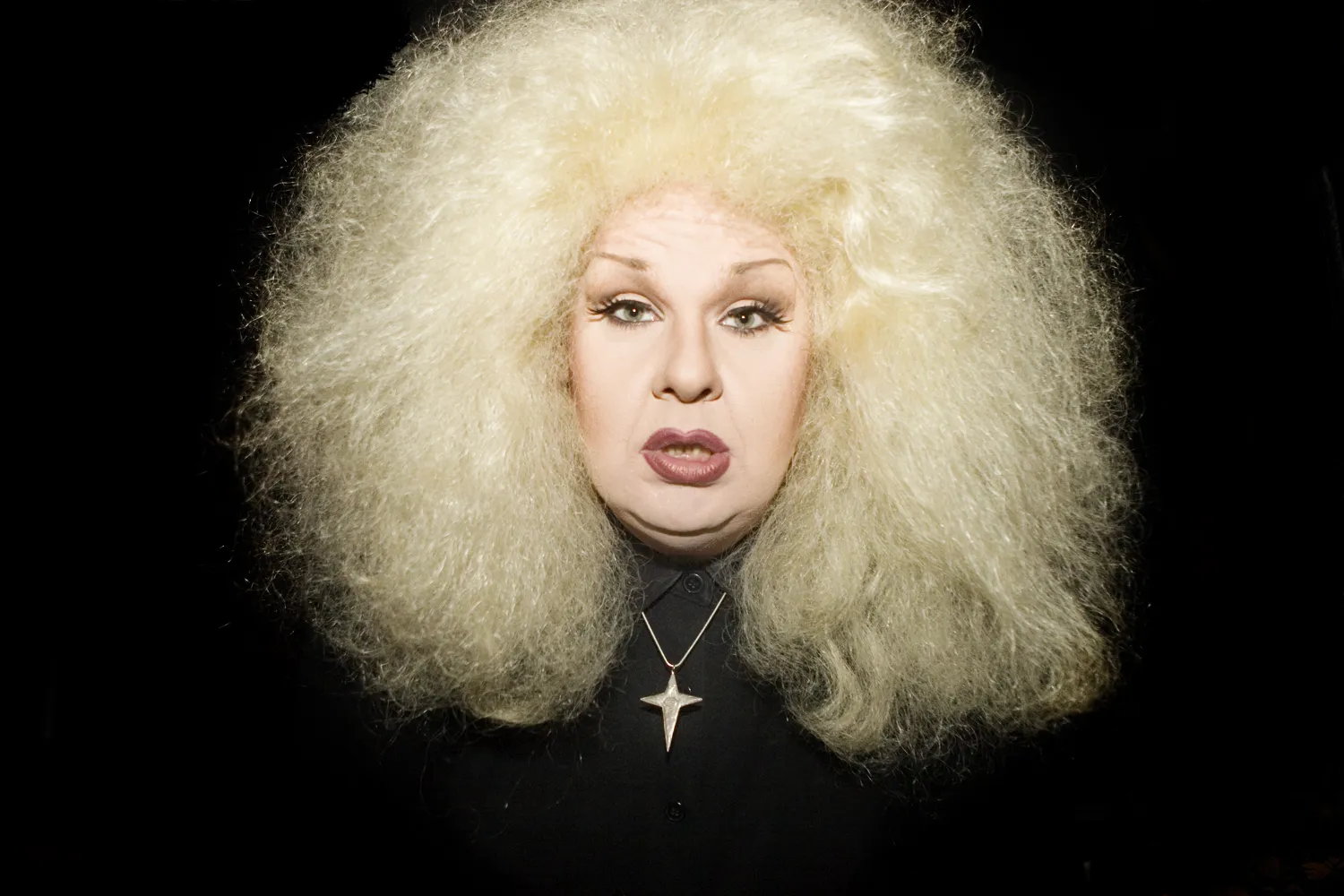 Portrait of a person with a large cloud of platinum-blond teased hair, dramatic eye makeup and dark lipstick, wearing a black shirt and a four-pointed star pendant, against a black background.